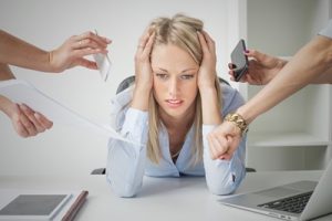 Lady with hands on head and elbows on a desk is looking stressed and overwhelmed as 4 arms try to get her to multitask. The Multitasking Myth. 