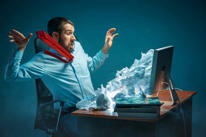 Young stressed handsome businessman working at desk in modern office shouting at laptop screen and being angry about e-mail spam. Collage with a mountain of crumpled paper. Business, internet concept (Young stressed handsome businessman working at desk